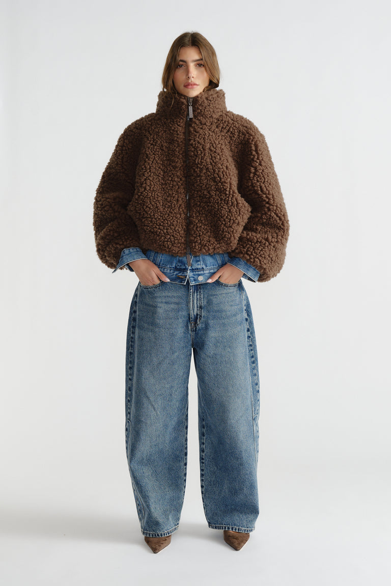 Woman wearing a cozy brown sherpa bomber jacket layered over a denim jacket paired with wide-leg blue jeans and pointed shoes