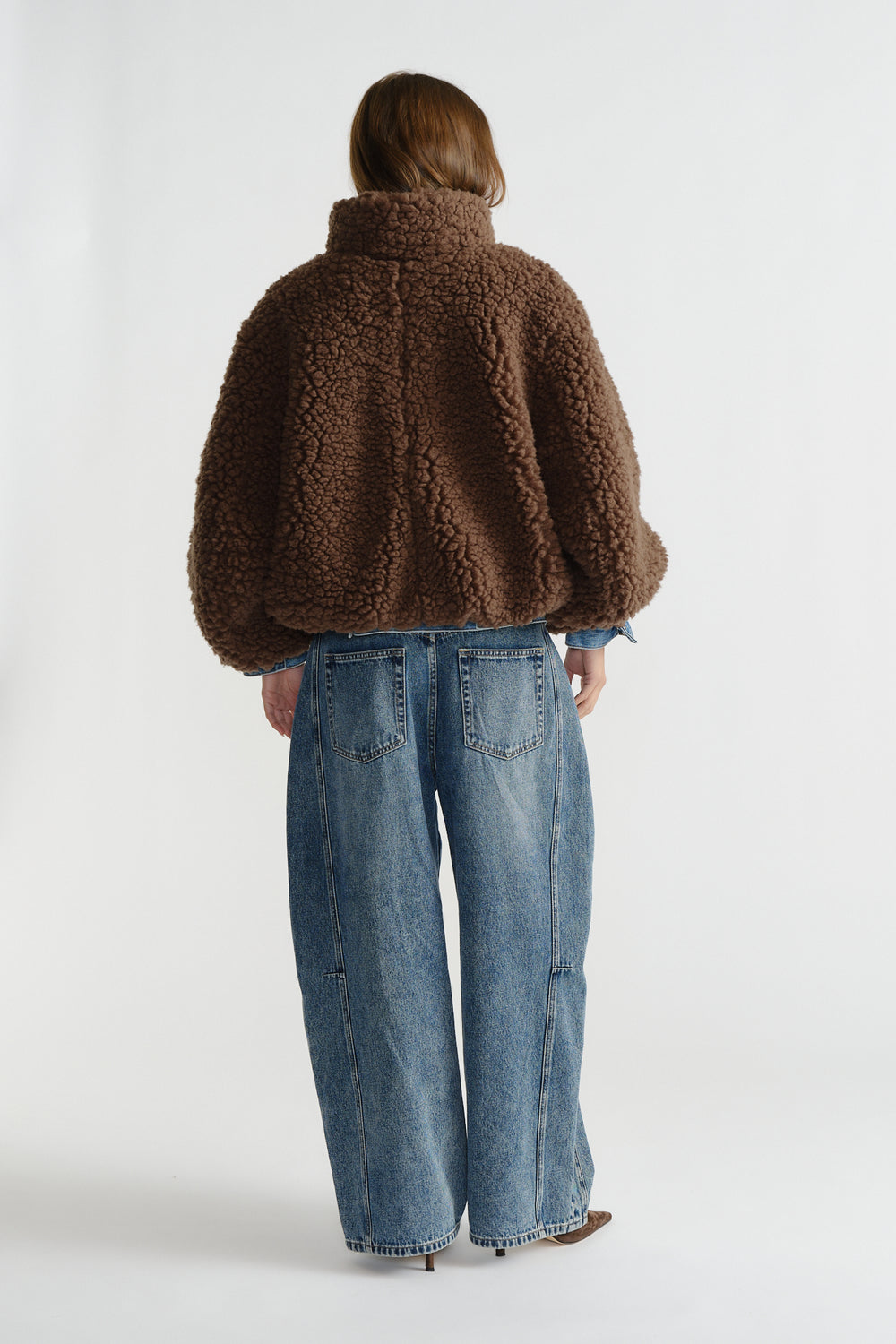 Back view of woman wearing a plush brown sherpa bomber jacket paired with relaxed wide-leg blue jeans.