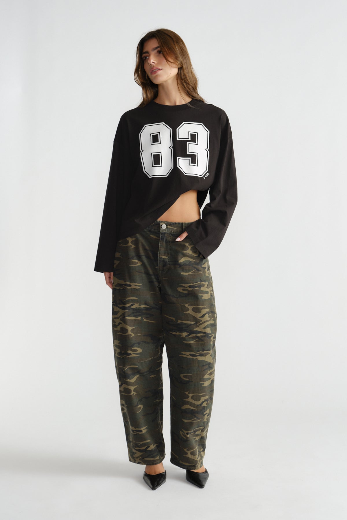Woman wearing a black long-sleeve top with bold number print paired with relaxed camo-patterned pants and black pointed shoes