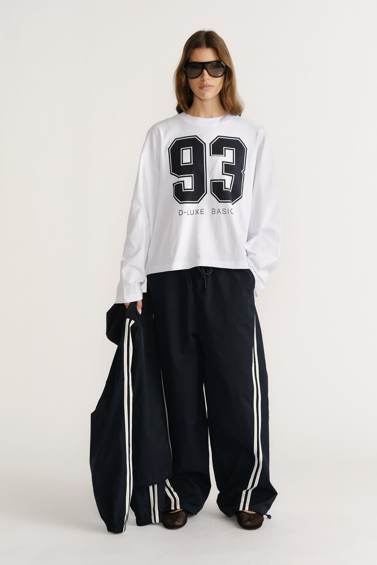 Woman wearing a white long sleeve graphic tee with black number print and loose black track pants with side stripes
