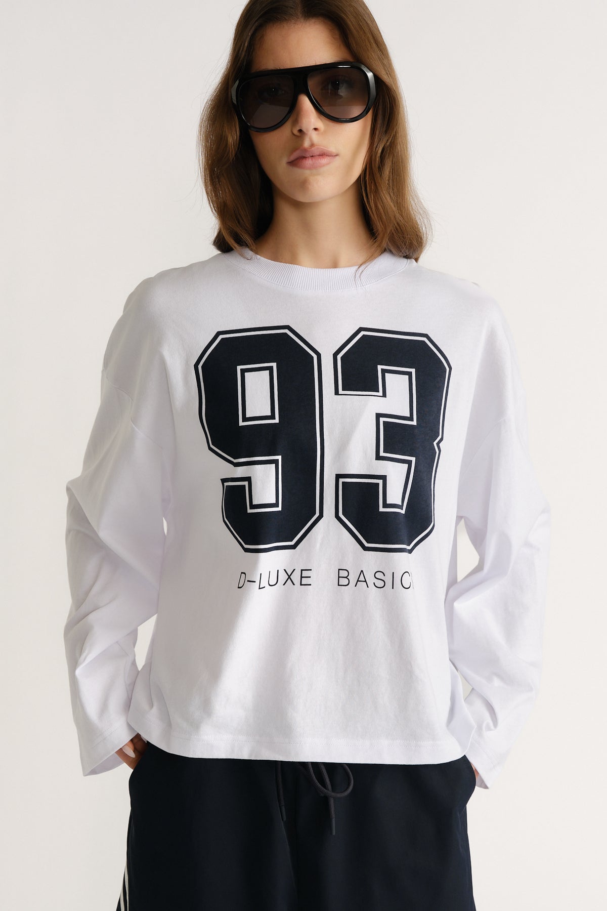Woman wearing a white long sleeve tee with bold black number print styled with oversized sunglasses and casual black pants