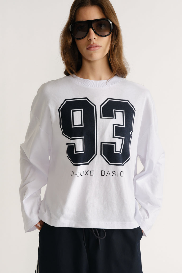 Woman wearing a white long sleeve tee with bold black number print styled with oversized sunglasses and casual black pants
