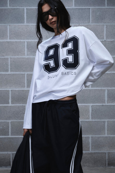 Woman wearing a white long sleeve 93 graphic tee with black track pants and side stripes