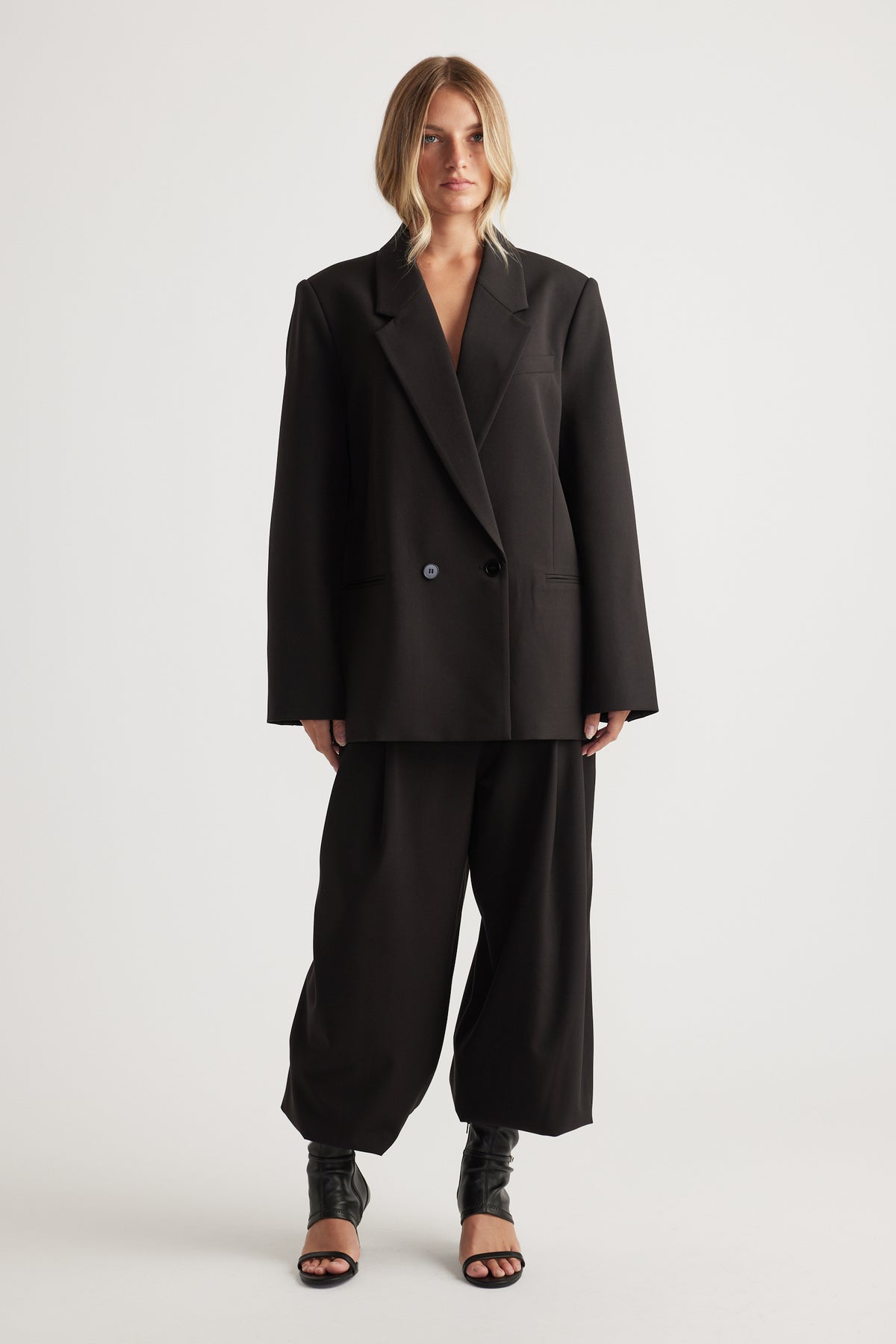Woman wearing an oversized black blazer and matching wide-leg pants paired with black open-toe heeled boots against a plain background
