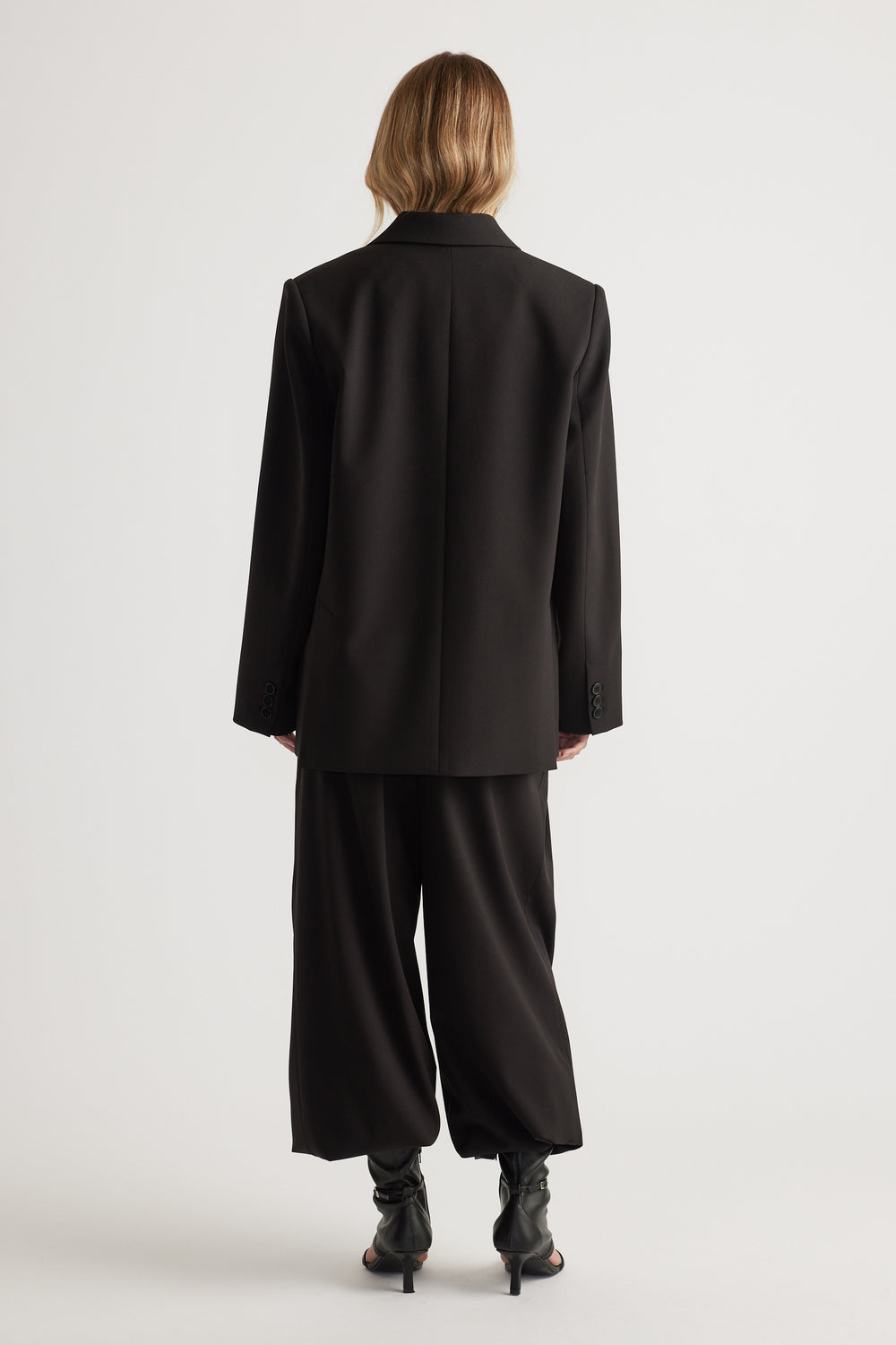 Model seen from behind wearing a black oversized blazer with relaxed trousers and heeled open-toe boots