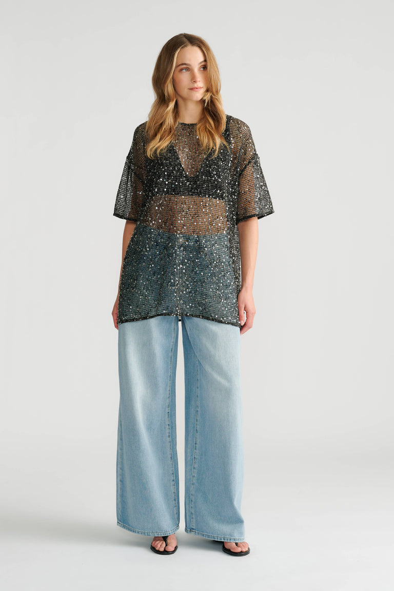 Woman wearing a sheer black sequin top layered over a black bralette paired with wide-leg light blue denim jeans.