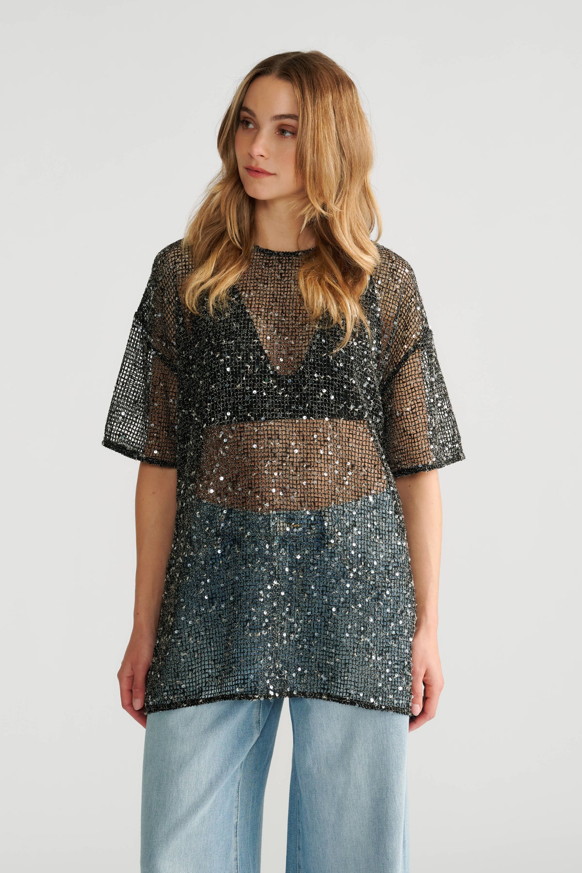 Woman wearing a sheer black sequin top layered over a black bralette, paired with loose light blue jeans.