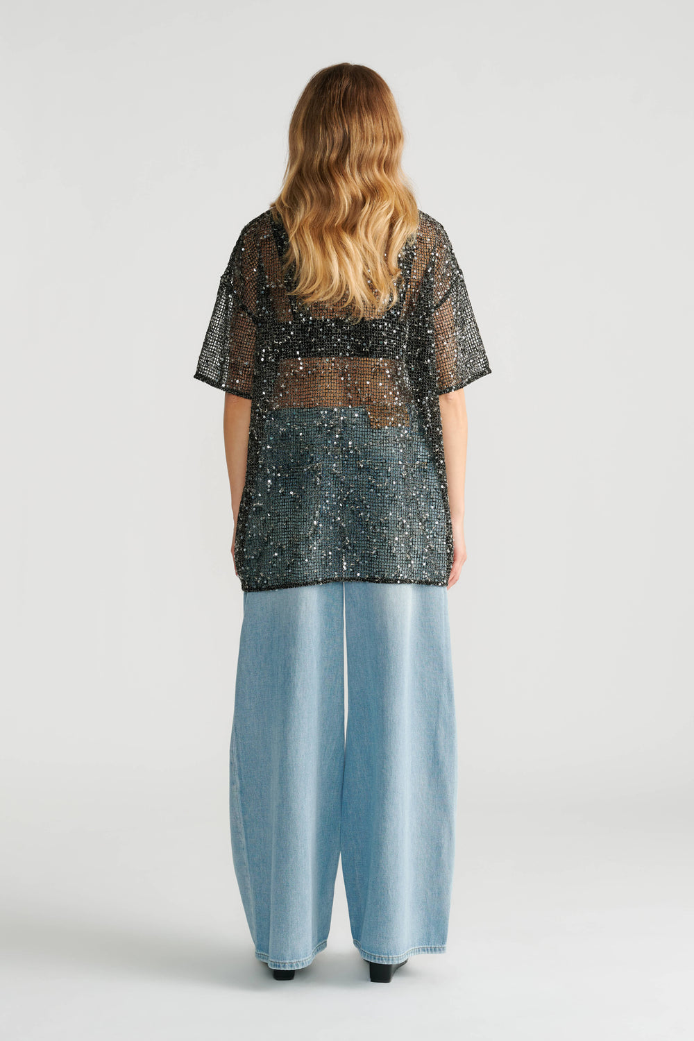 Woman wearing a black sequin mesh top layered over a black bralette paired with wide-leg light blue denim jeans.