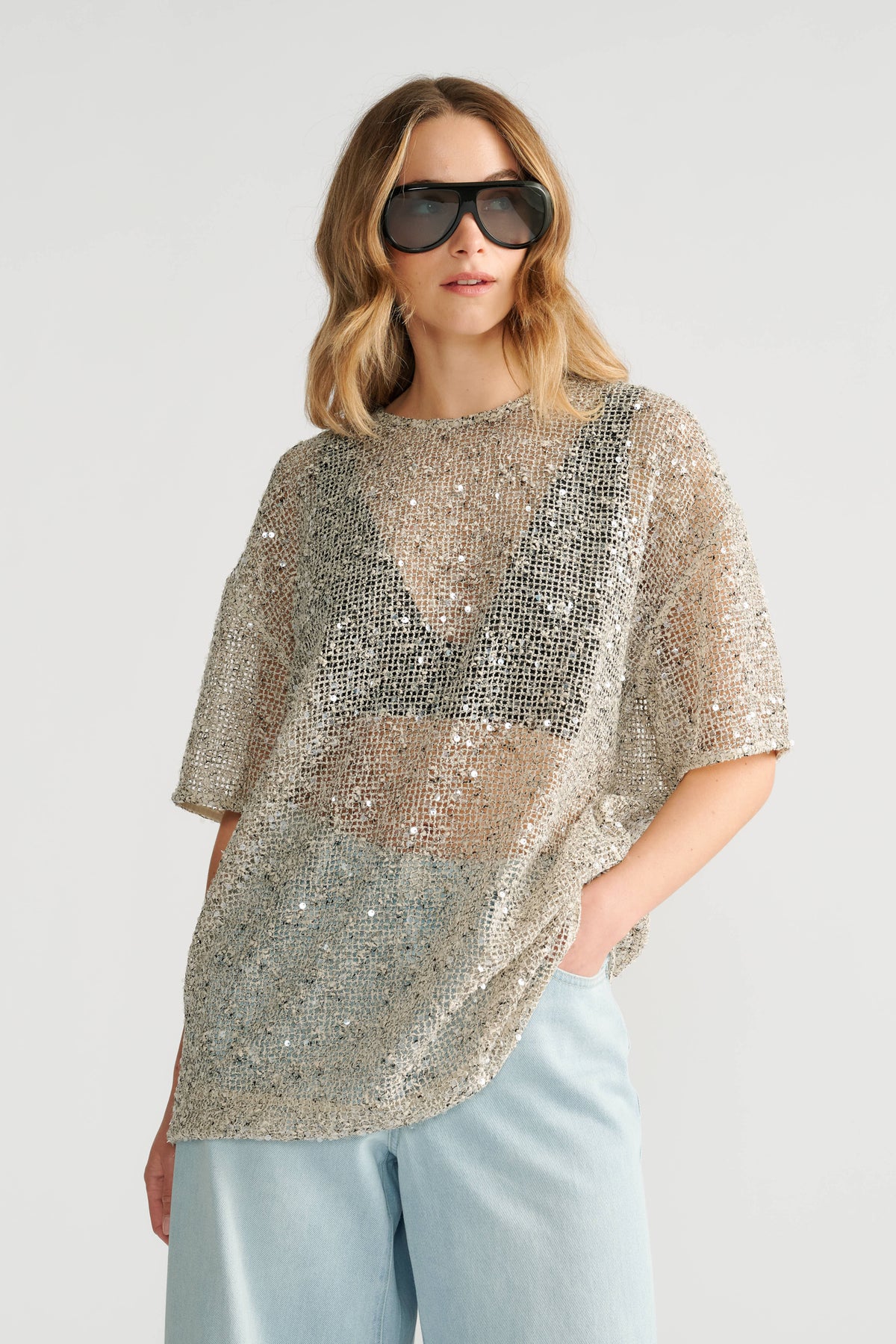 Woman wearing oversized silver sequin top over black bralette paired with light blue wide-leg denim jeans and sunglasses.
