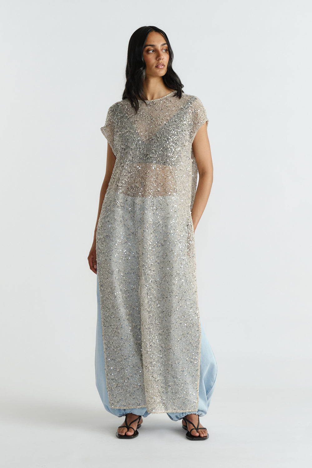 Model wearing a sleeveless long sequin dress layered over light blue wide-leg pants and black sandals.