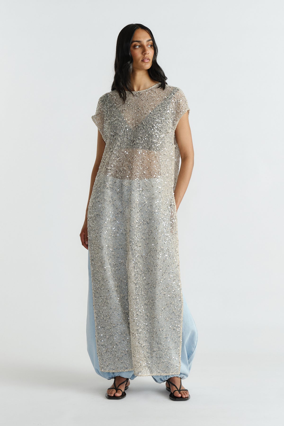 Model wearing a sleeveless long sequin dress layered over light blue wide-leg pants and black sandals.