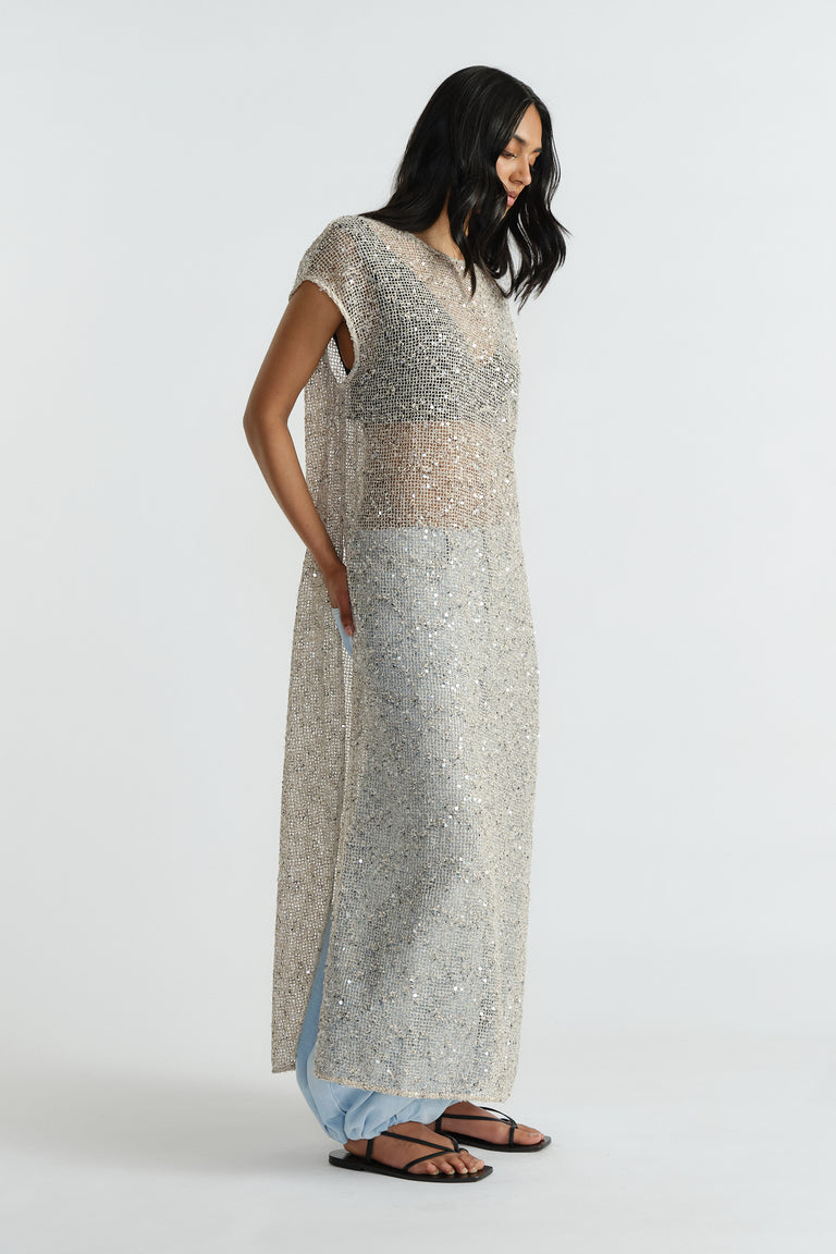 Side view of woman wearing a sleeveless, shimmering silver sequin dress layered over light blue pants with black sandals.