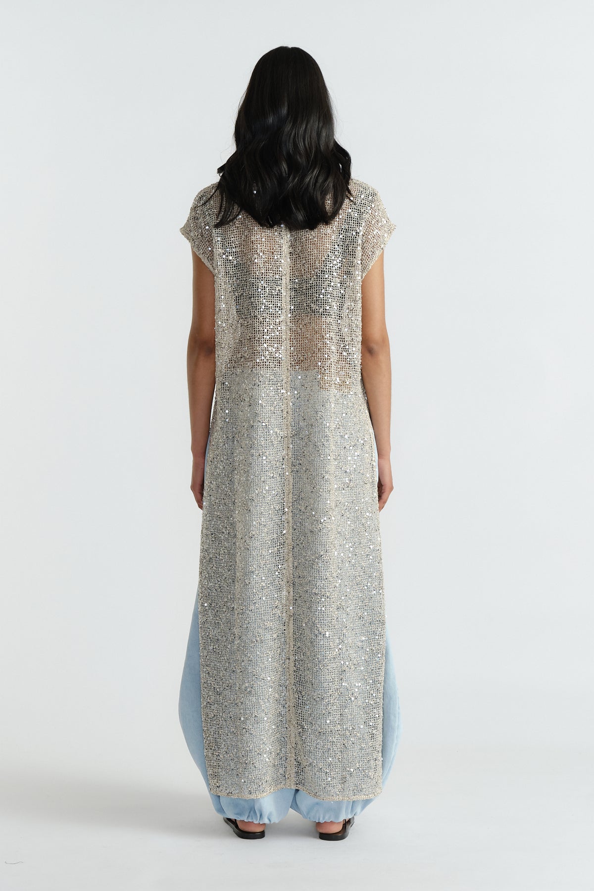 Back view of woman wearing a shimmering silver sequin dress layered over light blue wide-leg pants.