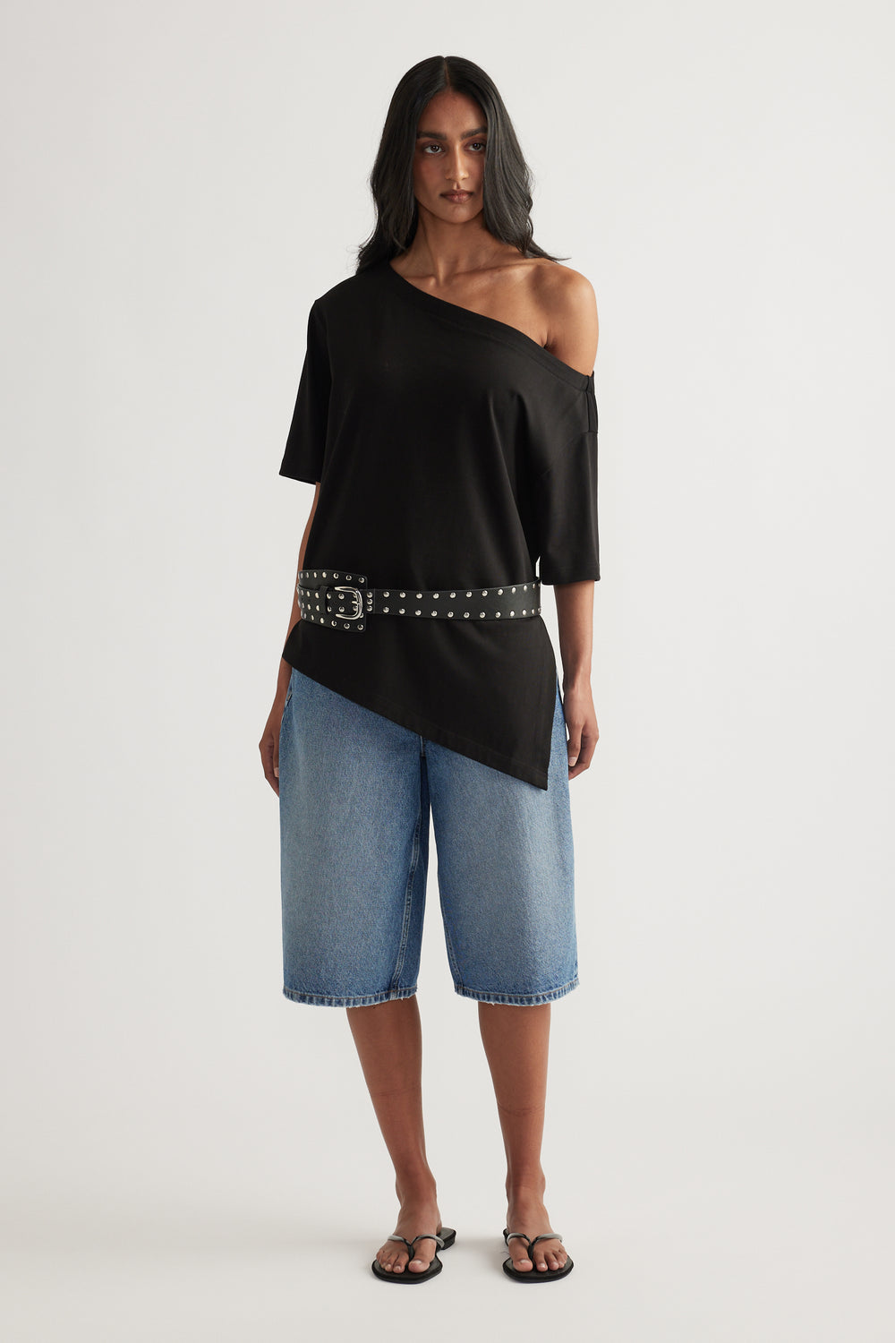 Woman wearing knee-length denim shorts with a black asymmetric top, studded belt and sandals