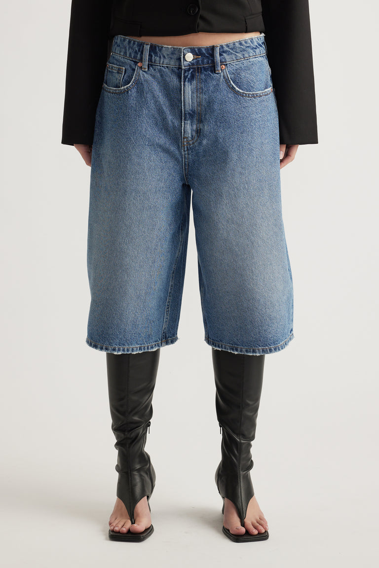 Woman wearing high waisted blue denim longline shorts with a black top and tall black boots