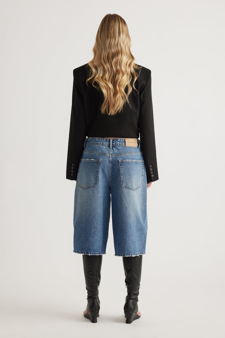 Back view of a woman wearing a black blazer, longline denim shorts and black knee-high boots