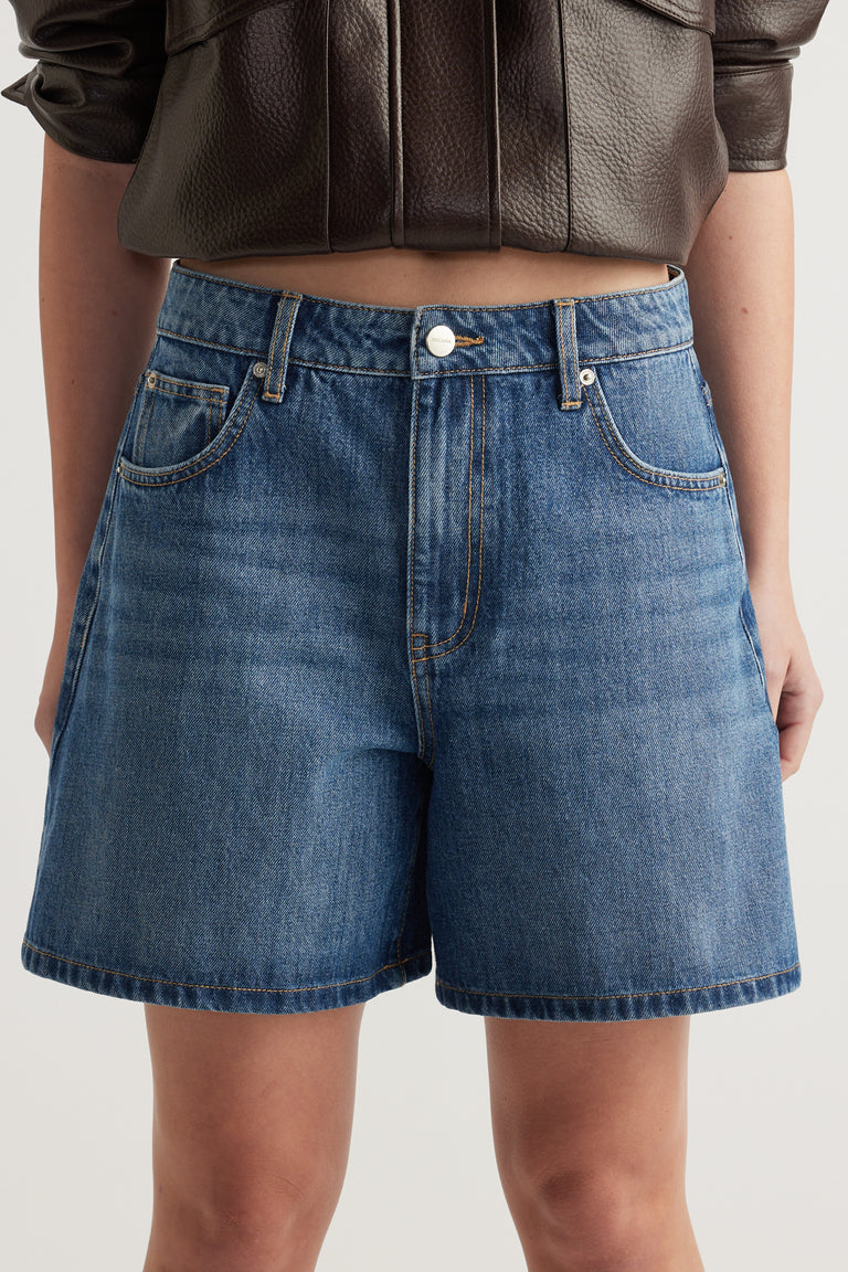 Woman wearing high rise blue A-line denim shorts with a button and zip fly and front pockets