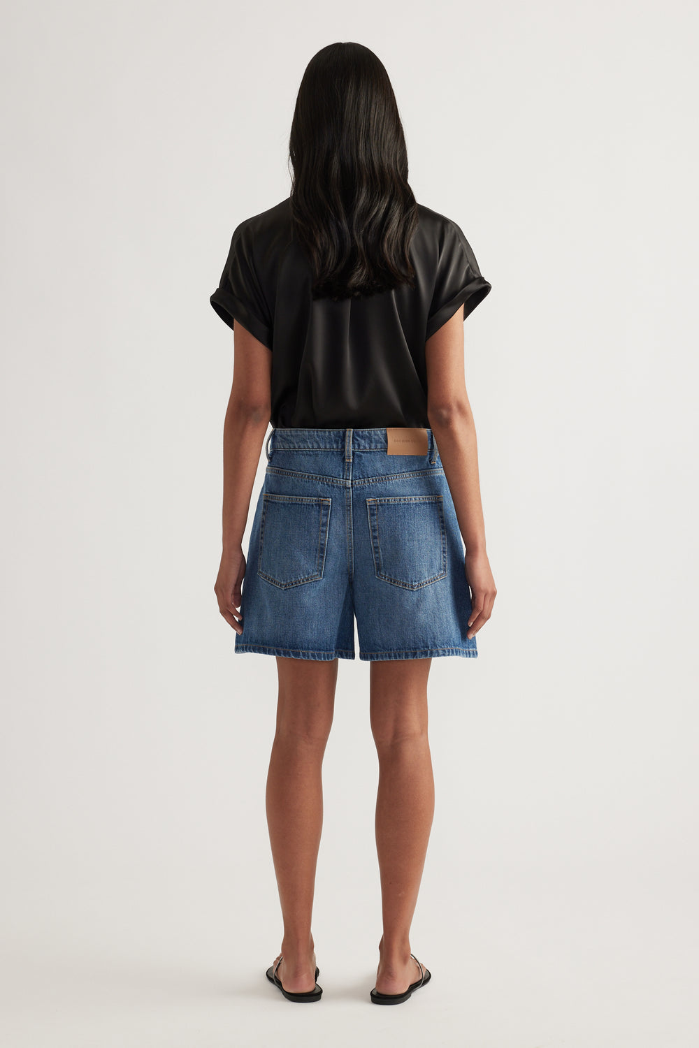 Back view of a woman wearing high rise blue denim A-line shorts with a black tee and sandals