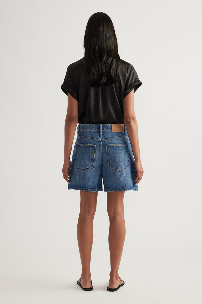 Back view of a woman wearing high rise blue denim A-line shorts with a black tee and sandals