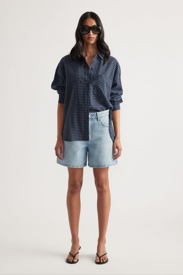 Woman wearing light blue high rise A-line denim shorts paired with an oversized navy checkered shirt and black sandals