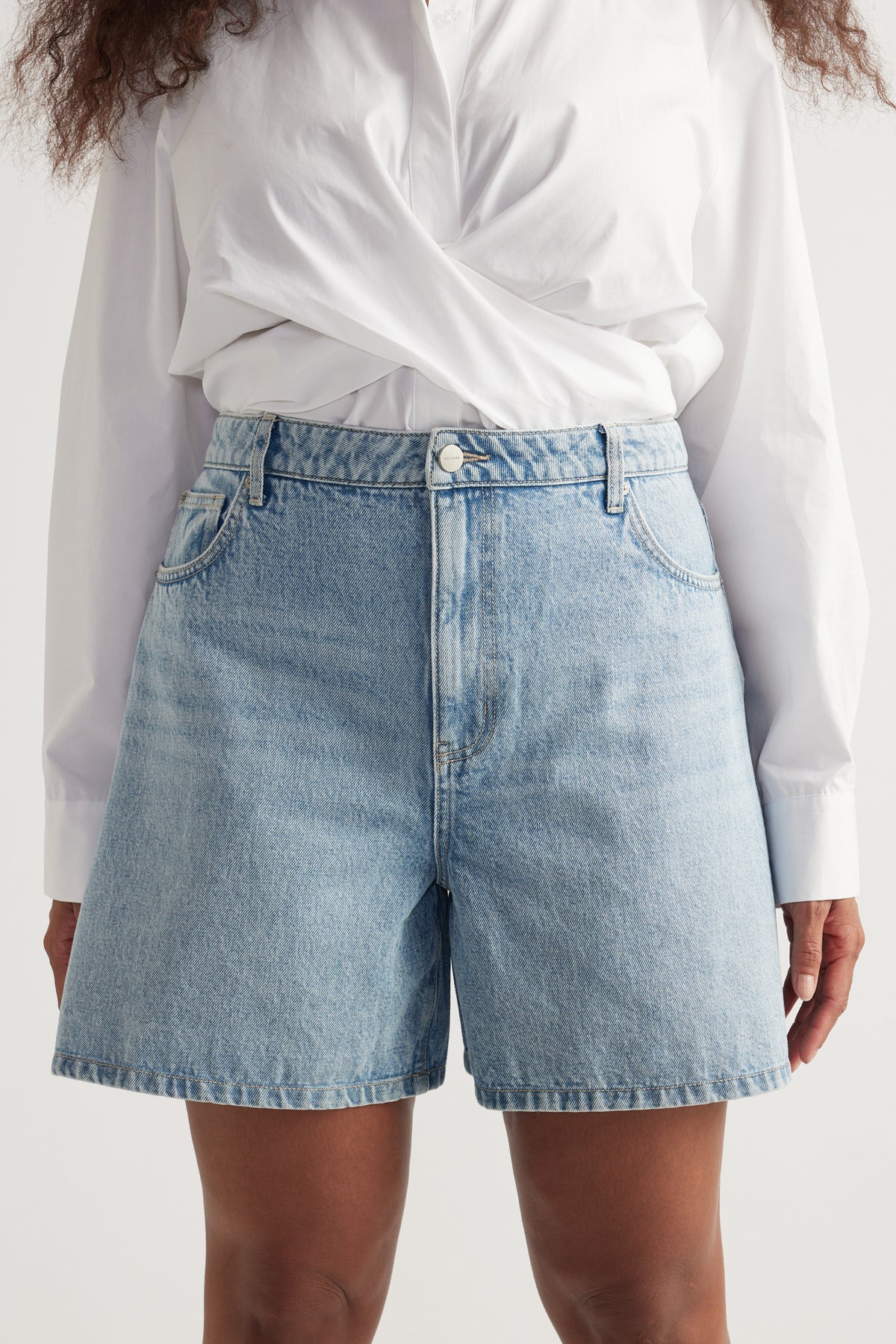 Woman wearing light wash high rise A-line denim shorts paired with a crisp white long sleeve shirt
