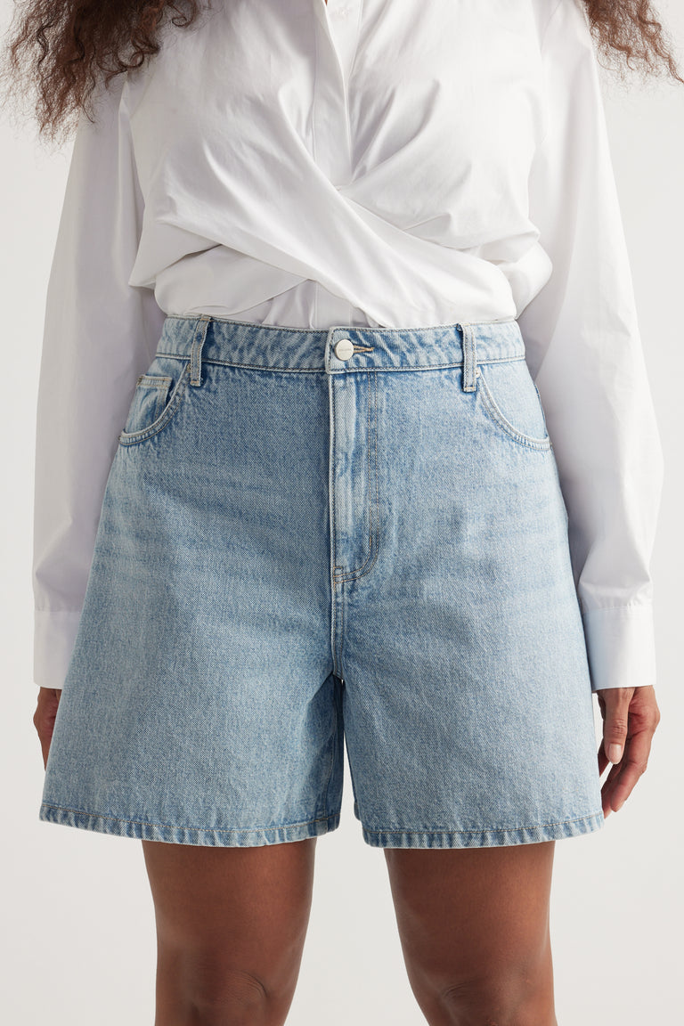 Woman wearing light wash high rise A-line denim shorts paired with a crisp white long sleeve shirt