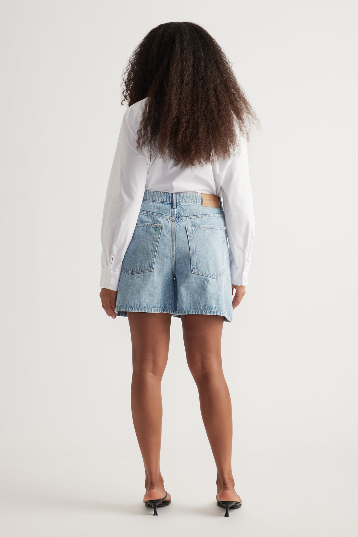 Rear view of woman wearing light wash high rise A-line denim shorts with back pockets and white long sleeve shirt