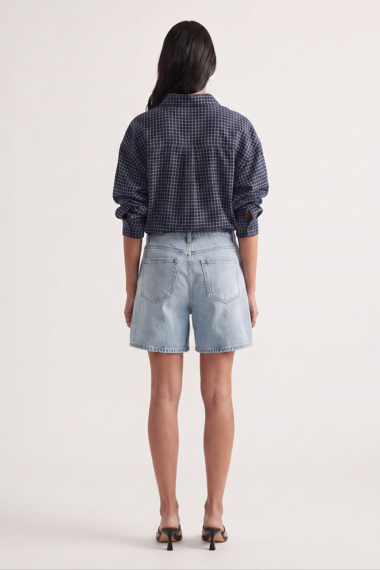 Rear view of a woman wearing light wash high-rise denim shorts with a navy checkered long sleeve shirt and heels
