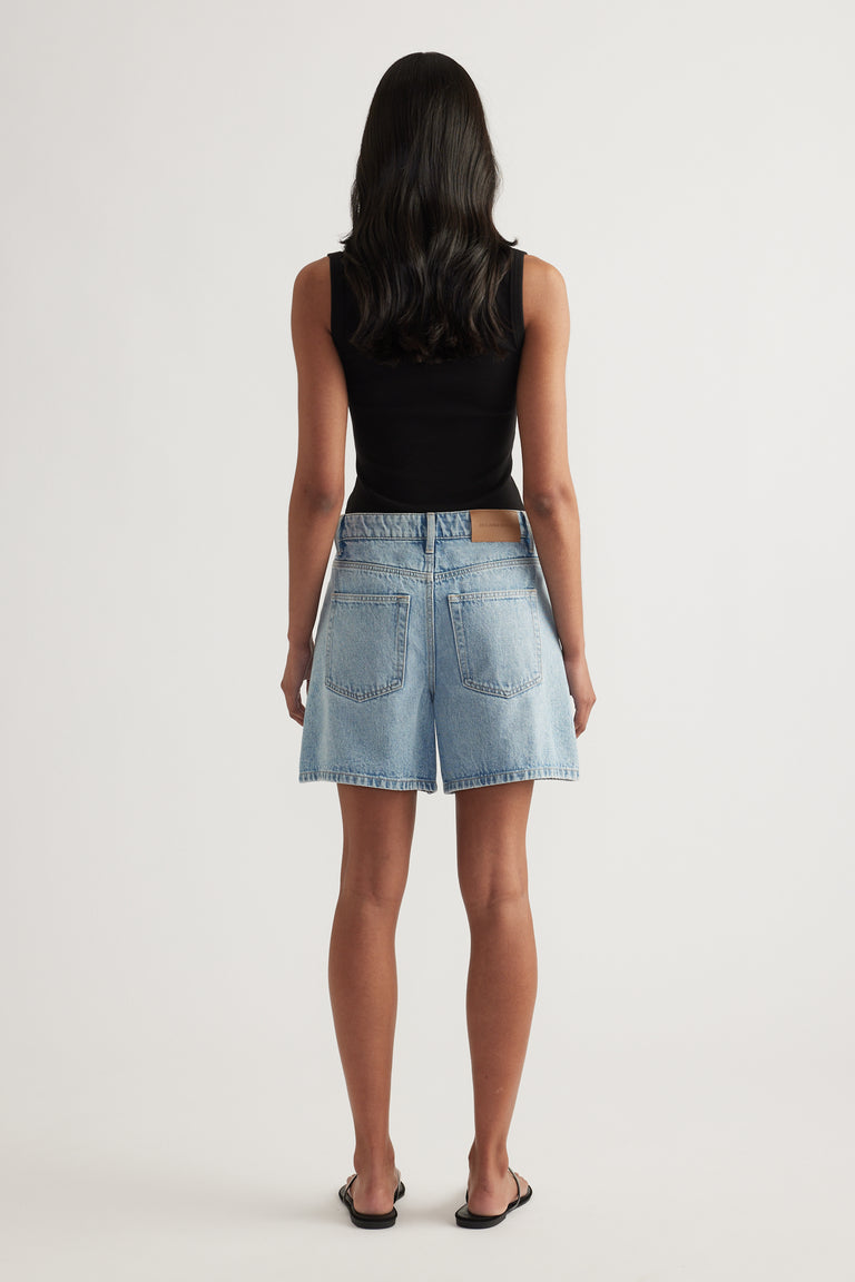 Back view of a woman wearing high rise denim A-line shorts with a black top and sandals