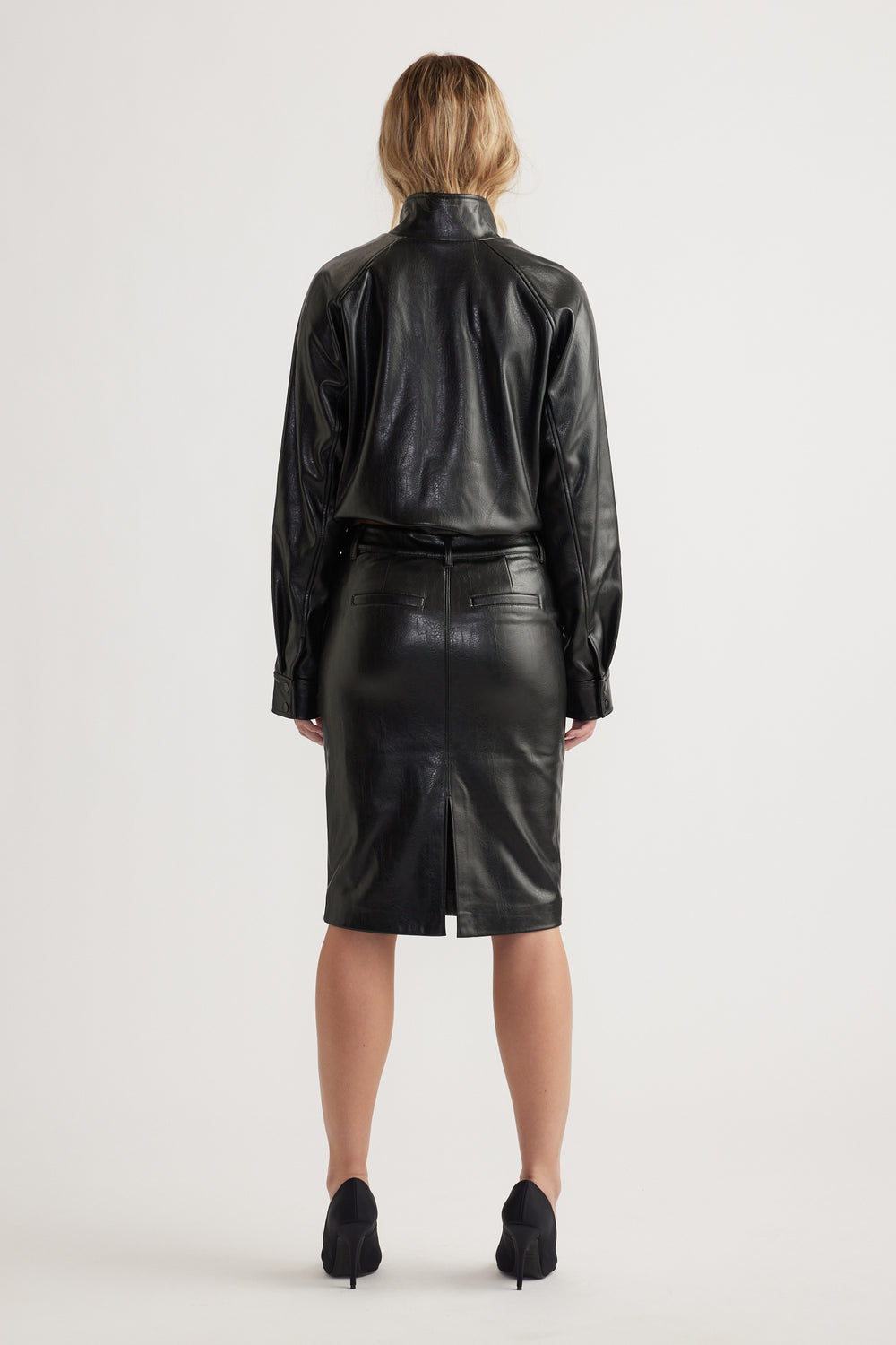 Woman wearing black faux leather shirt and high-waist pencil skirt with back slit and belted waist seen from behind