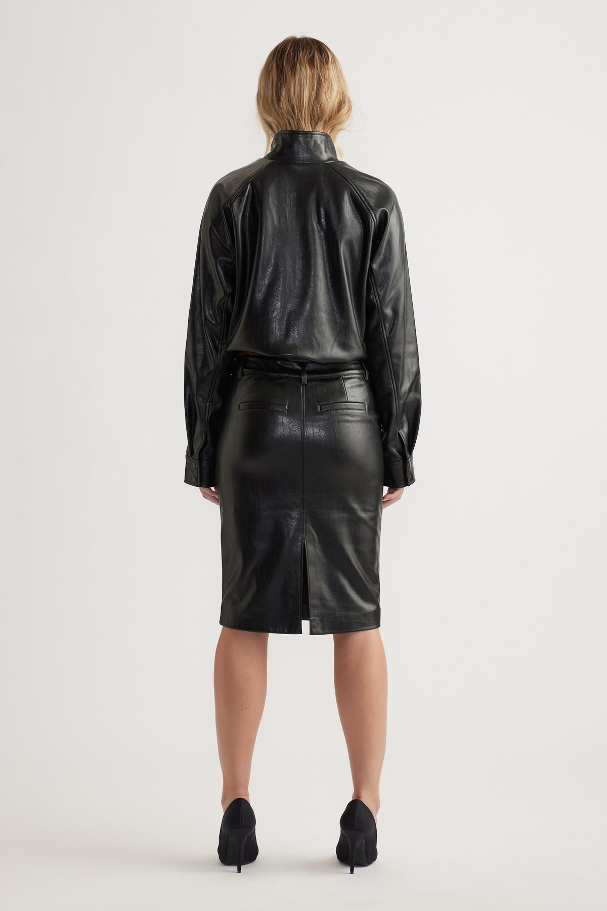 Woman wearing black faux leather shirt and high-waist pencil skirt with back slit and belted waist seen from behind
