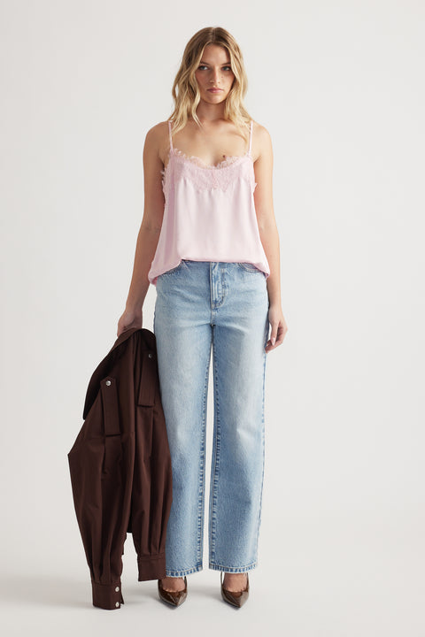 Woman wearing a soft pink lace-trimmed cami with light blue high-waisted jeans, holding a dark brown jacket and pointed heels