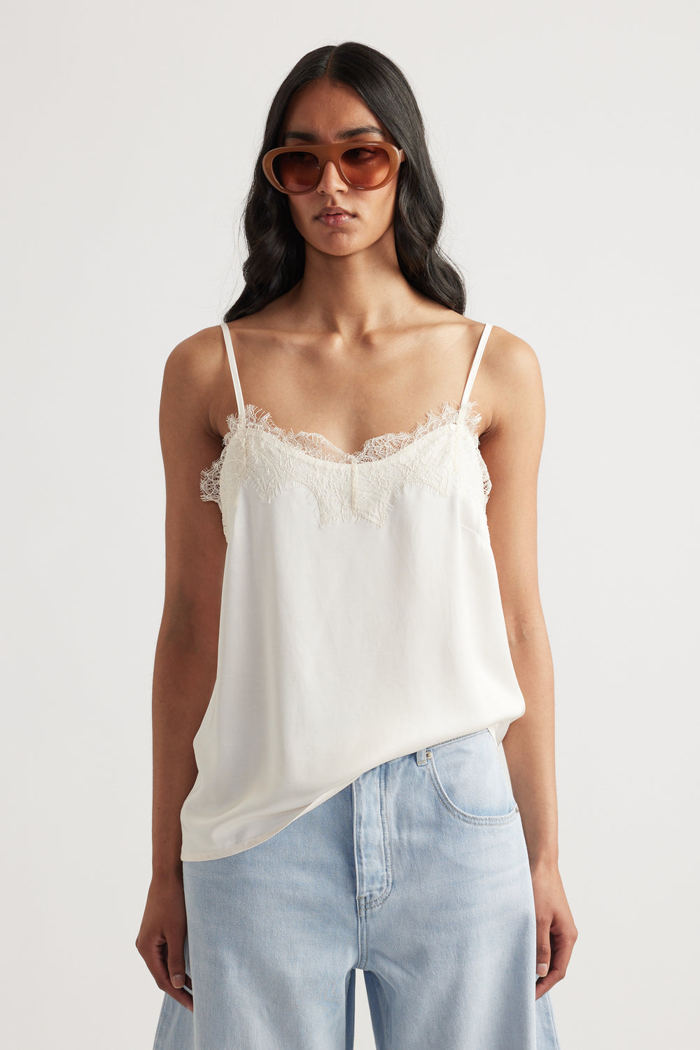 Woman wearing a delicate ivory lace-trimmed cami with thin straps paired with light blue relaxed jeans and oversized sunglasses