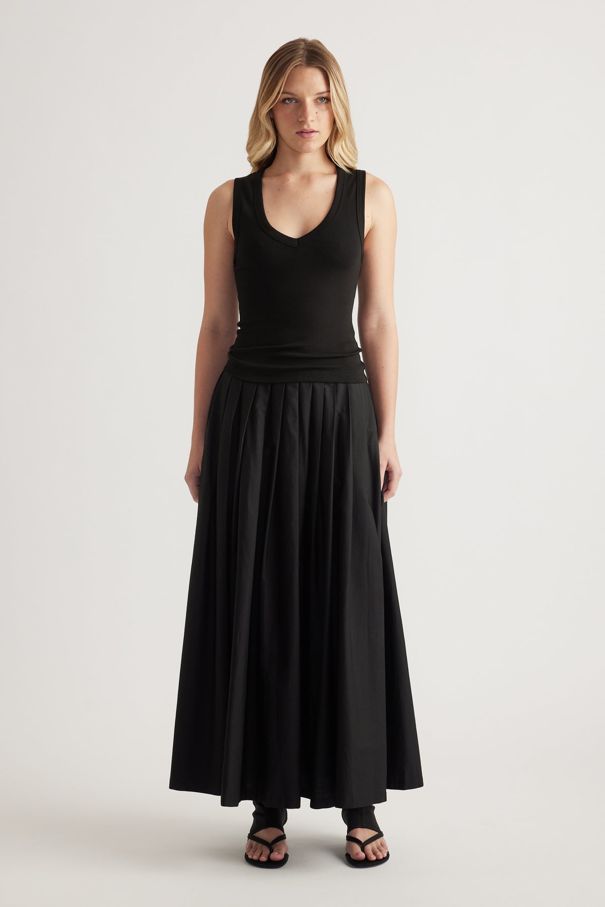 Model wearing a black pleated ankle length skirt with a sleeveless top and sandals