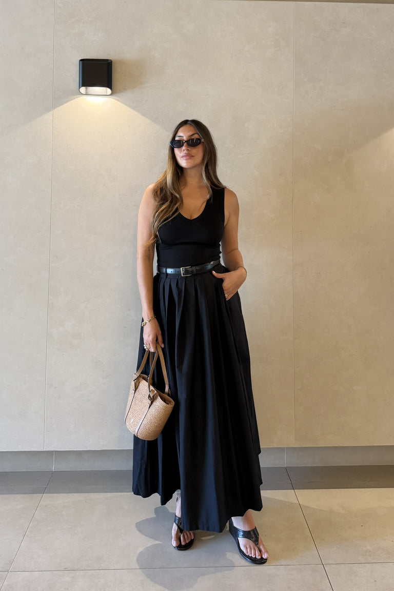 Woman wearing a black pleated ankle length skirt with a belt and woven handbag