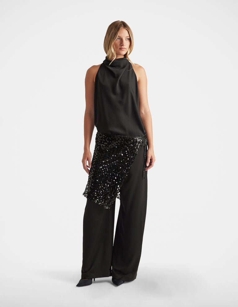 Woman wearing a sleeveless black draped top paired with wide-leg pants featuring a sequined asymmetric overlay.