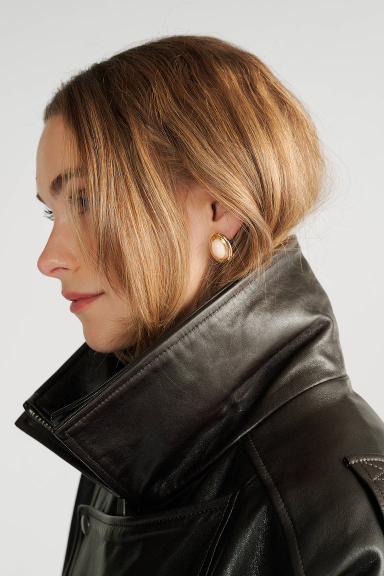 Woman with blonde hair wearing elegant gold-toned stud earrings and a sleek black leather jacket with a high collar.