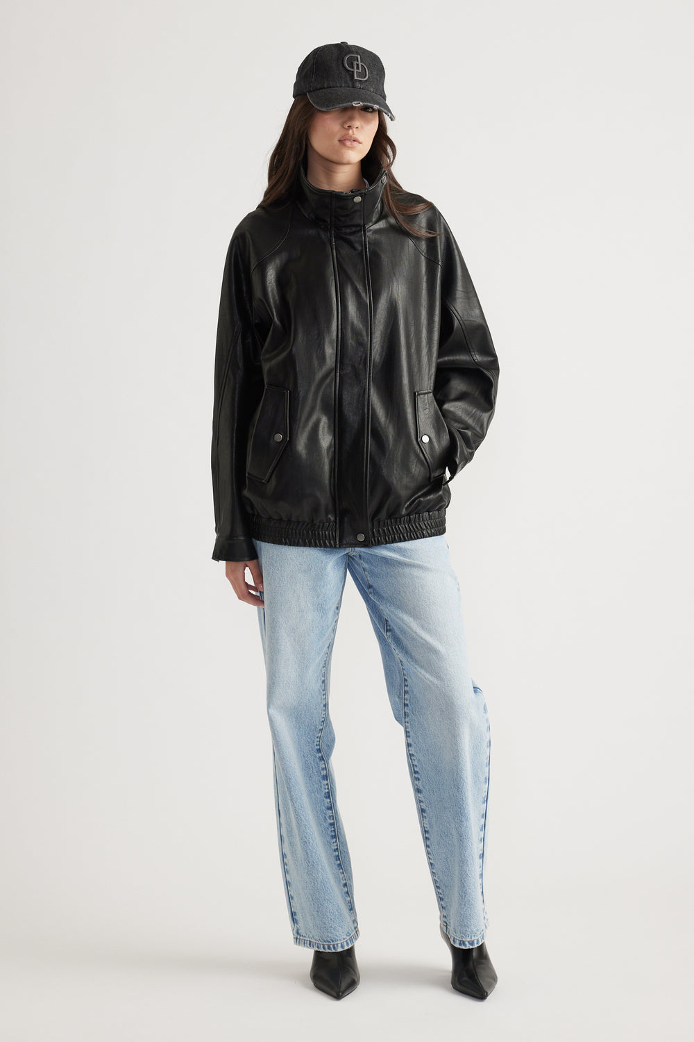 Woman wearing black faux leather jacket with snap pockets, light blue jeans, and black denim baseball cap for casual style.