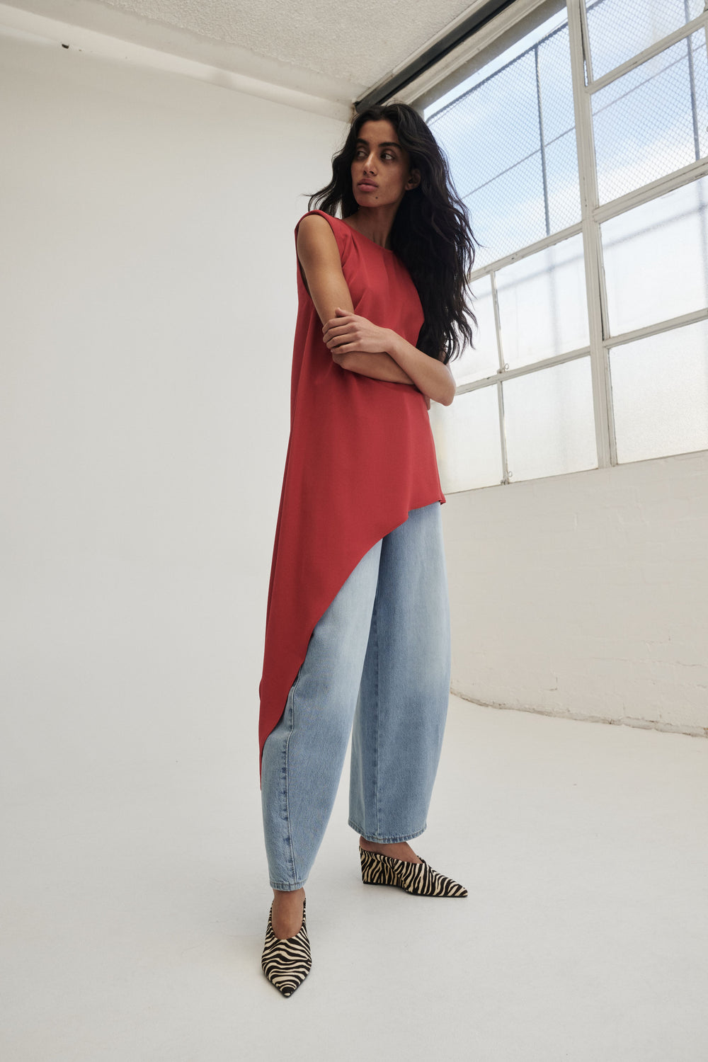 Woman wearing a red sleeveless draped top with padded shoulders, light blue jeans and zebra print flats