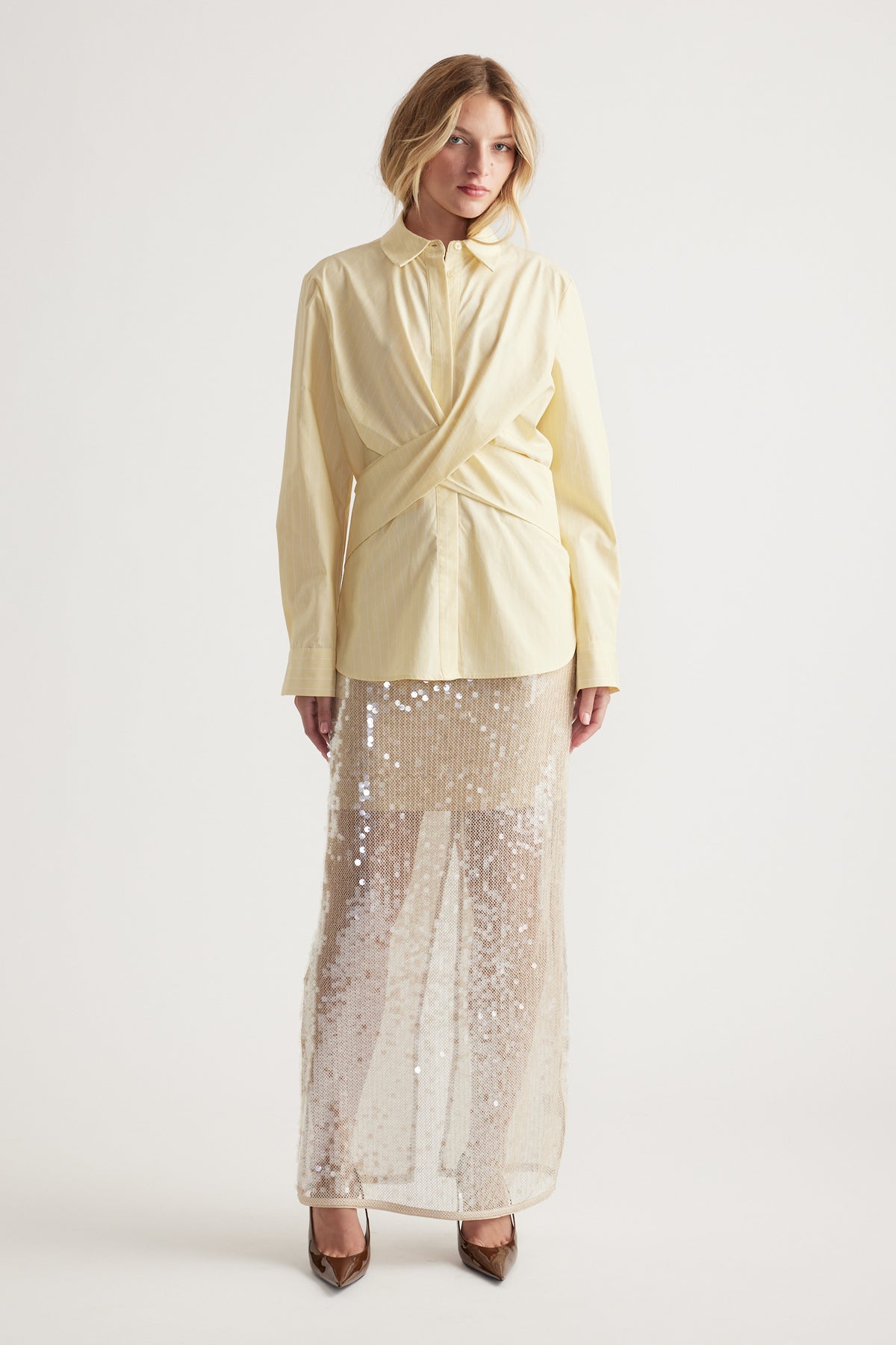 Woman wearing a pale yellow wrap-style shirt paired with a long, sheer sequin skirt and pointed heels