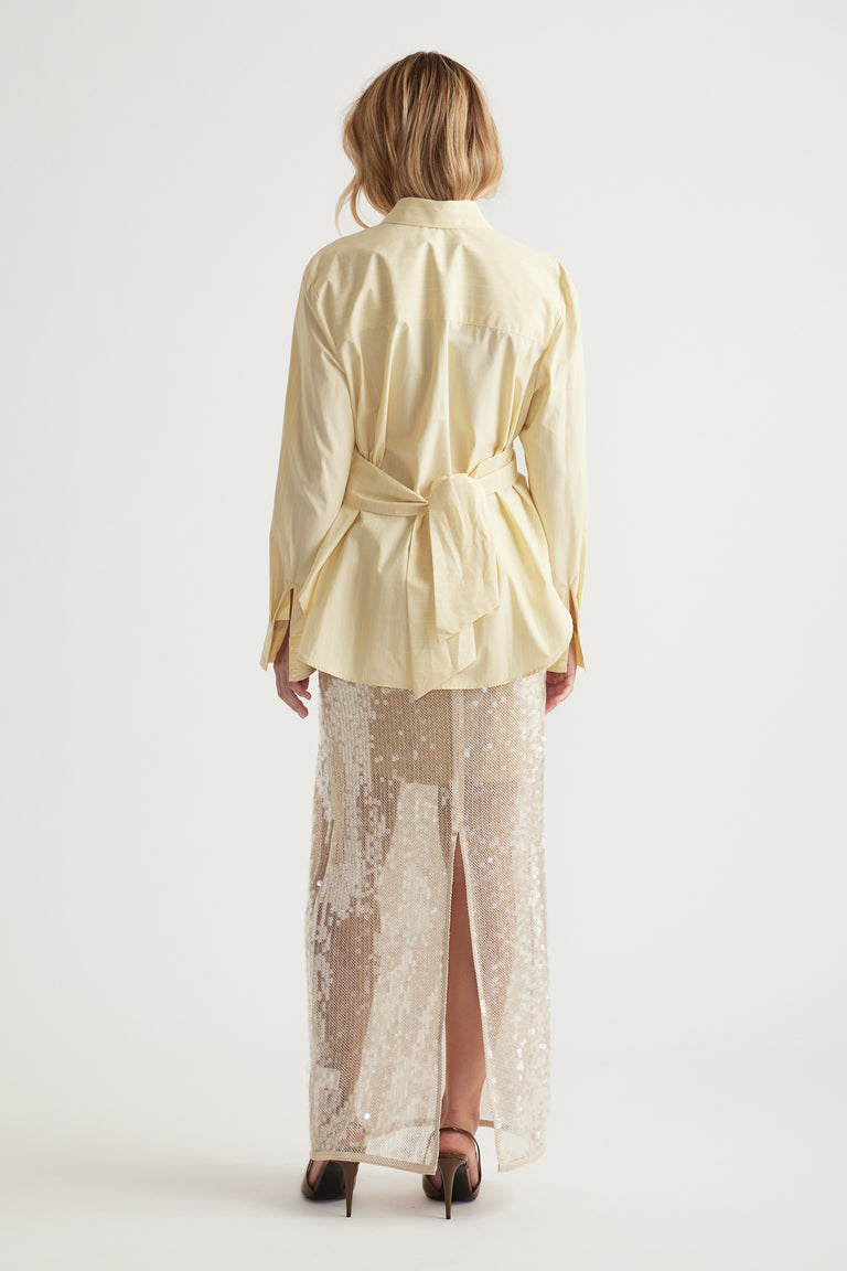 Woman wearing a loose pale yellow tie-back shirt paired with a long sequined skirt and black heels, back view