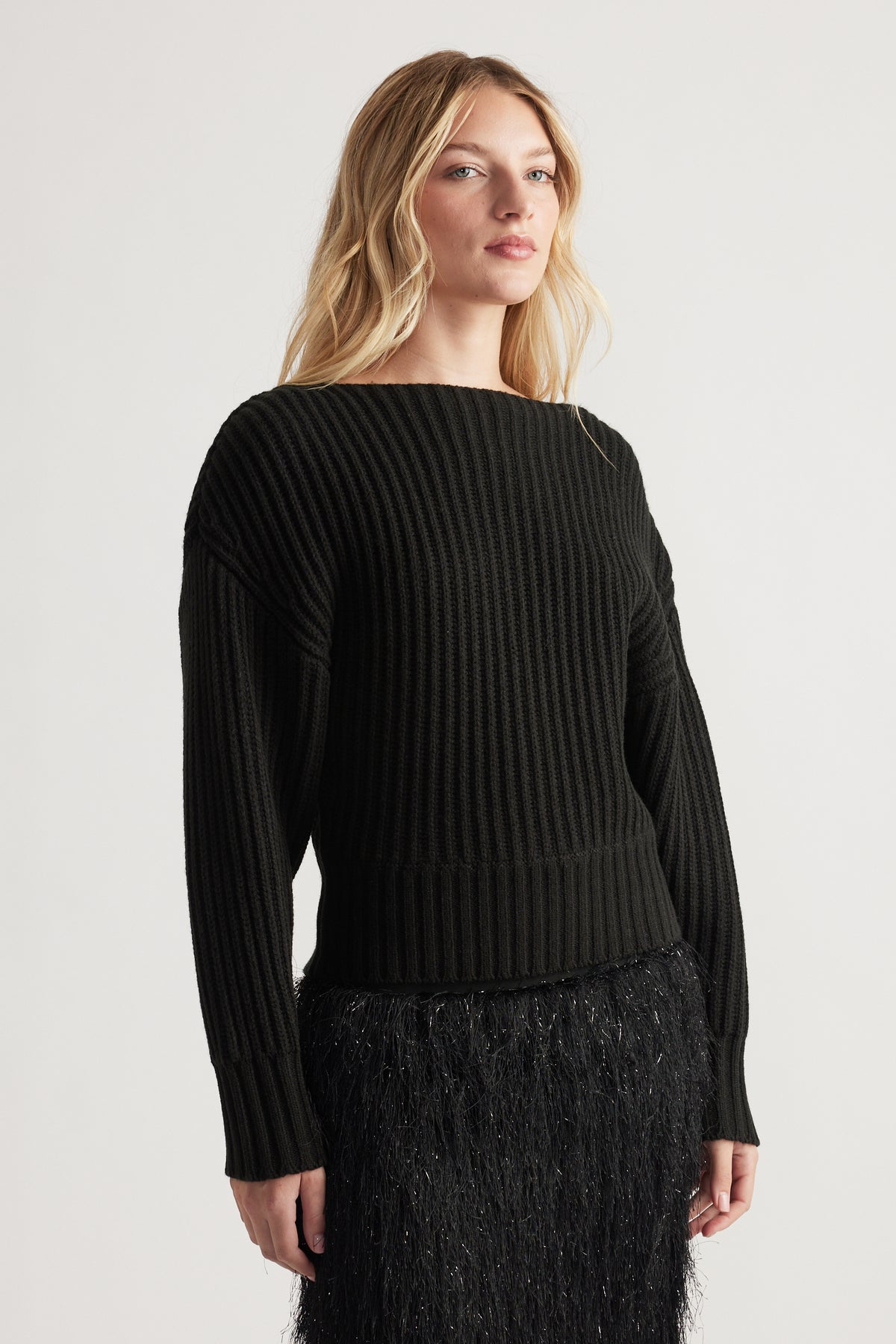 Woman wearing a black ribbed knit jumper with a drop shoulder design paired with a textured black skirt against a plain background.