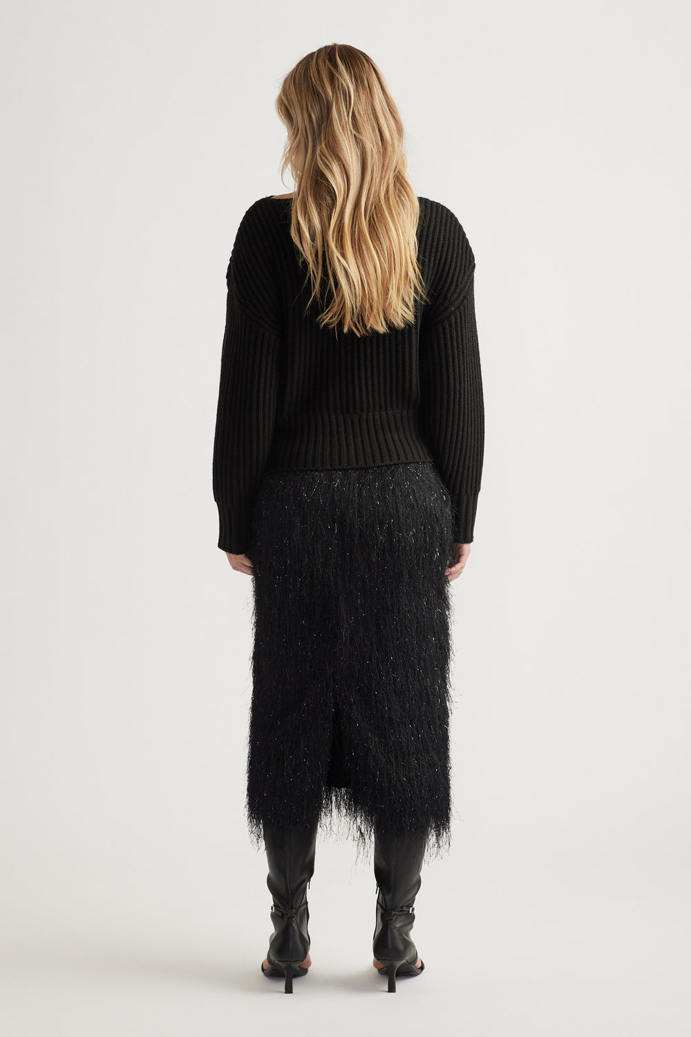 Woman seen from behind wearing a black ribbed knit jumper with drop shoulders and a textured midi skirt with black boots.