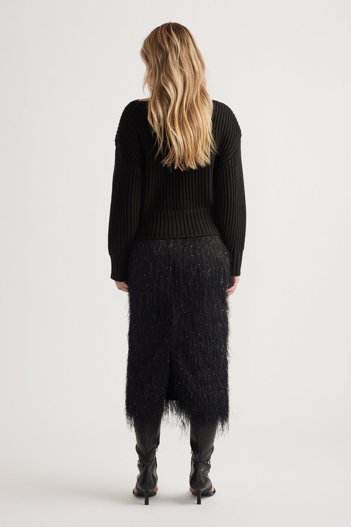 Woman seen from behind wearing a black ribbed knit jumper with drop shoulders and a textured midi skirt with black boots.