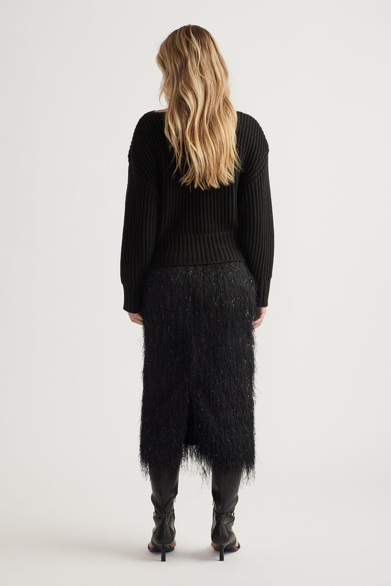 Woman seen from behind wearing a black ribbed knit jumper with drop shoulders and a textured midi skirt with black boots.