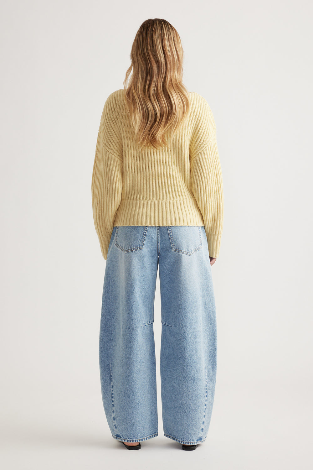 Back view of woman wearing a pale yellow drop shoulder knit jumper with relaxed wide-leg light denim jeans