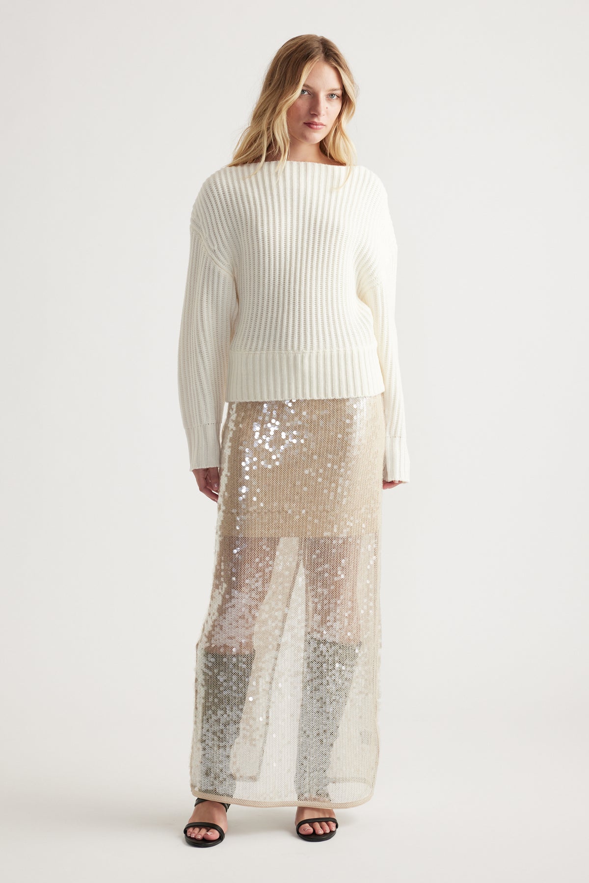 Woman wearing an off-white ribbed drop shoulder knit jumper paired with a sheer sequin maxi skirt and black sandals.