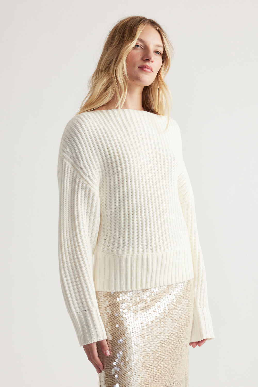 Woman wearing a cream knit jumper with drop shoulders paired with a shimmering sequin skirt against a plain background