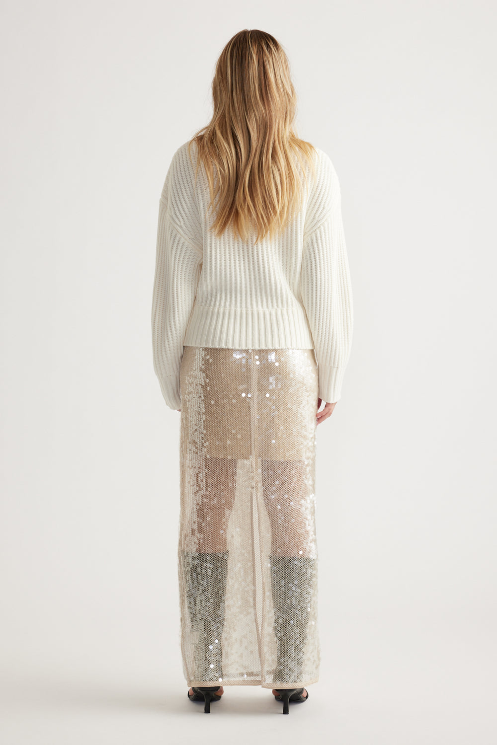 Woman wearing an off-white ribbed knit jumper paired with a shimmering sequin maxi skirt and black heels, shown from behind.