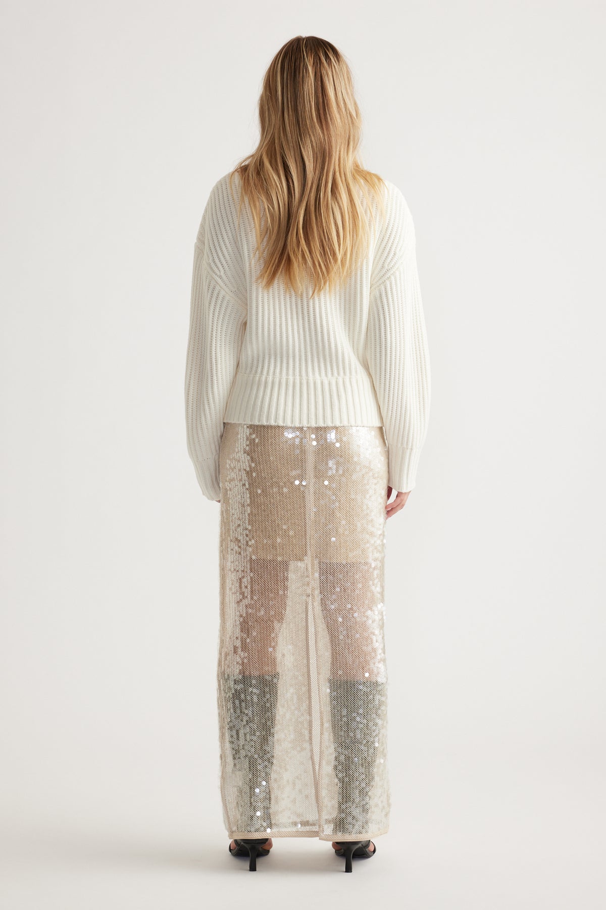 Woman wearing an off-white ribbed knit jumper paired with a shimmering sequin maxi skirt and black heels, shown from behind.